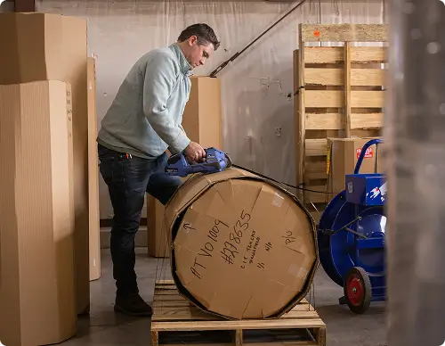Warehouse worker using a strapping tool to secure a large cardboard-wrapped fuel tank on a pallet