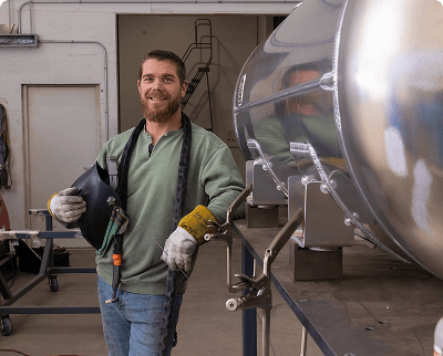 Smiling worker in a workshop wearing work gloves and holding welding gear with a large polished aluminum fuel tank in the background