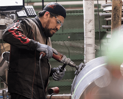 Technician using a power tool on a large metal fuel tank cylinder while wearing safety glasses and gloves