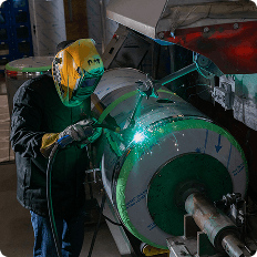 Welder in a yellow protective helmet welding a large cylindrical metal tank in an industrial workshop