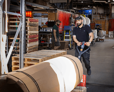 Warehouse worker using a pallet jack to move large cardboard-wrapped fuel tanks in an industrial warehouse