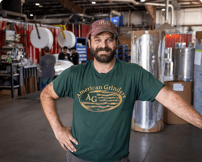 Smiling man wearing an American Grinder t-shirt and cap standing in an industrial warehouse with aluminum tanks in the background