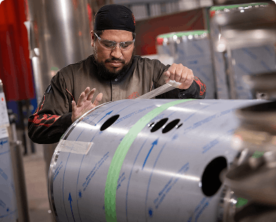 Worker examining and inspecting a large aluminum fuel tank with template markings in a manufacturing facility
