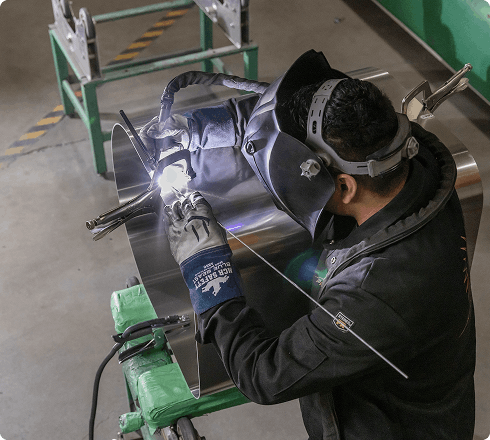 Overhead view of a welder working on an aluminum fuel tank component on a workbench in a manufacturing workshop