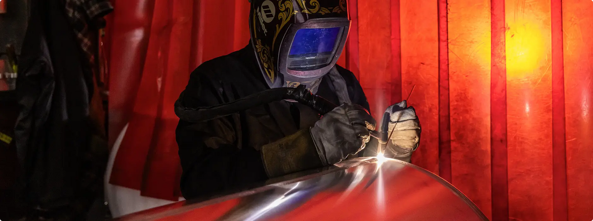 Welder in protective gear welding a polished aluminum fuel tank with a vivid red background