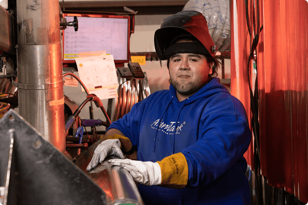 Alumitank worker wearing a blue Alumitank hoodie, work gloves, and a welding helmet looking at the camera in an industrial setting