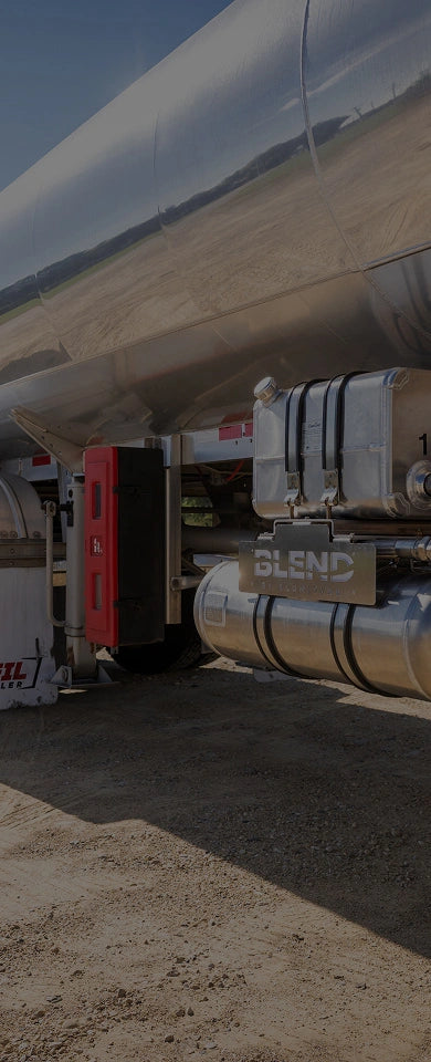Side view of a commercial tanker truck showing polished aluminum fuel tanks and fuel system components on a dirt road