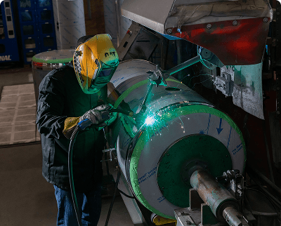 Welder in a yellow protective helmet welding a large cylindrical metal tank in an industrial repair shop